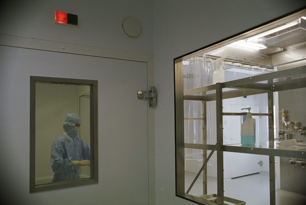 Cleanroom worker in protective gown and mask seen through a door window; a metal shelving unit with spray bottles visible through a second window.